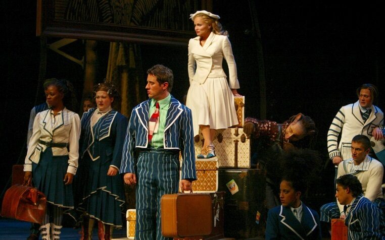 glinda in white shiz outfit on top of luggage with fellow shiz students