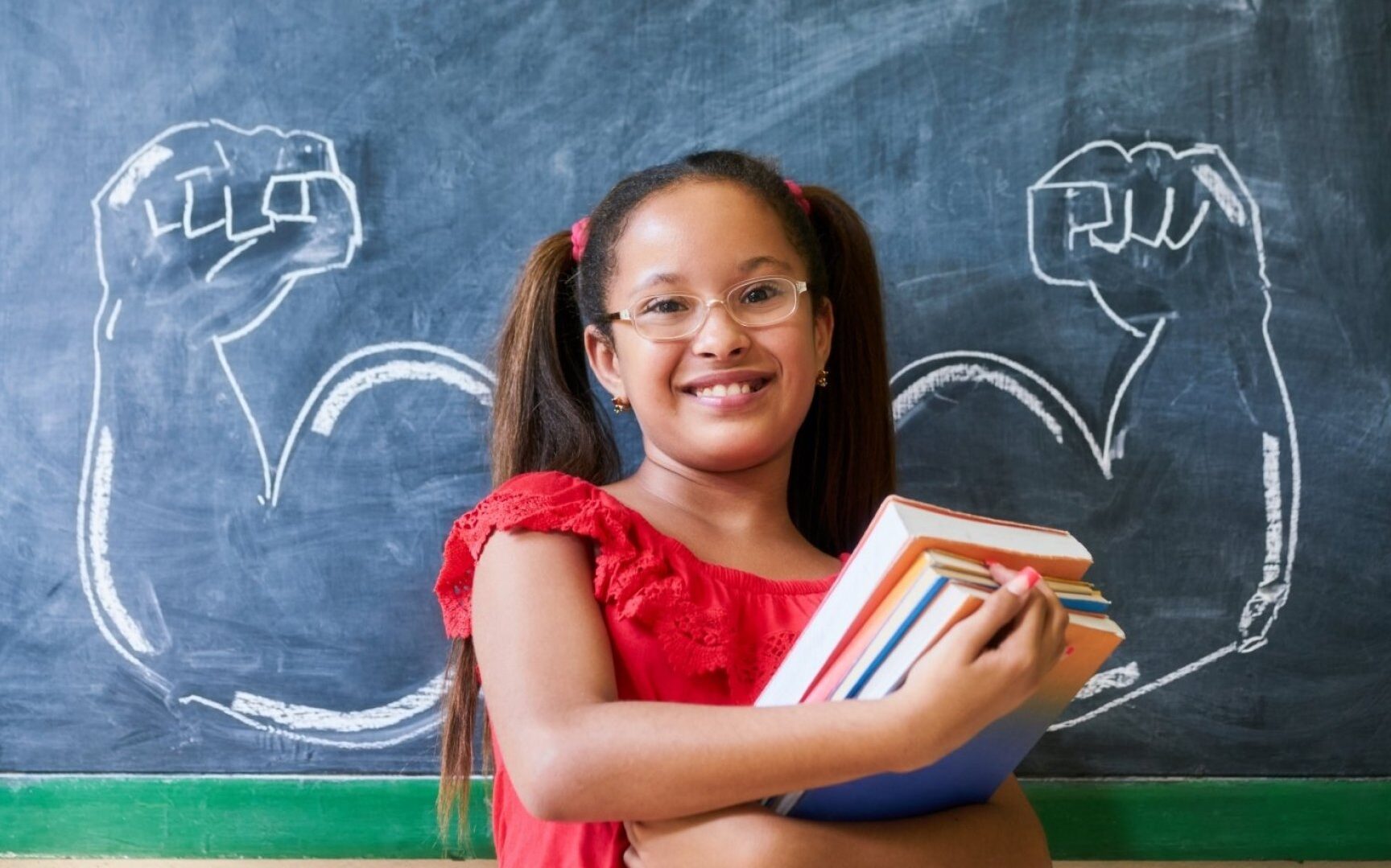 kid holding books in front of chalkboard with strong arms drawn on it